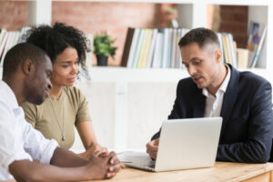 A couple is sitting at a table, discussing how to rent your house with a man in a suit who is showing them information on a laptop. 