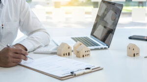Person working at a desk with a clipboard, small wooden house models, and a laptop displaying charts.