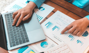 Person analyzing charts and graphs on printed documents while working on a laptop.