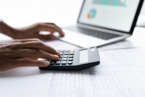 A person is using a calculator with one hand while typing on a laptop with the other.