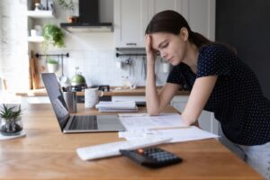 A young woman standing in her kitchen, leaning on the counter with her hand on her head, looking stressed. She is surrounded by bills, a calculator, and a laptop, as she ponders how much should I charge for rent. 