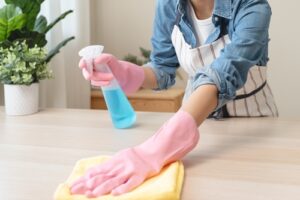 A person wearing pink gloves is cleaning a surface with a yellow cloth and holding a spray bottle of blue cleaning solution.