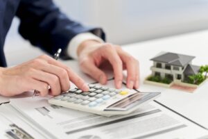 Person using a calculator with documents on a desk and a small house model in the background.