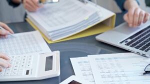 Spreadsheets, a binder, and a laptop being reviewed by two people at a desk. 