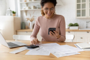 A woman sitting at a kitchen table using a smartphone and calculator, reviewing documents and smiling.